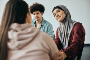A woman wearing a headscarf offers a comforting smile and supportive gesture to another person during a group discussion.