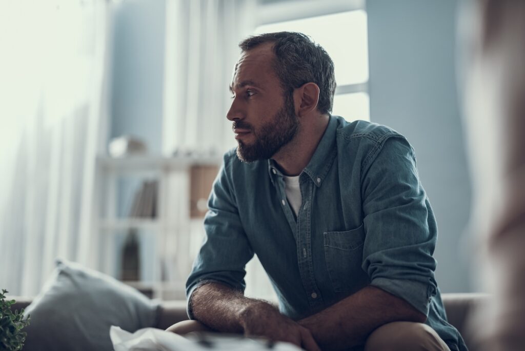 Man sitting indoors leans forward with a thoughtful expression, appearing deep in conversation or reflection.