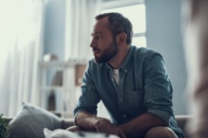 Man sitting indoors leans forward with a thoughtful expression, appearing deep in conversation or reflection.