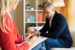 Man sitting on a couch with his hands clasped looks down thoughtfully while a therapist in a red jacket takes notes on a clipboard.