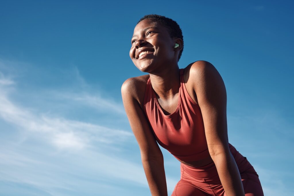 A smiling woman in athletic wear looks upward after exercising outdoors under a bright blue sky.