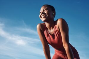 A smiling woman in athletic wear looks upward after exercising outdoors under a bright blue sky.