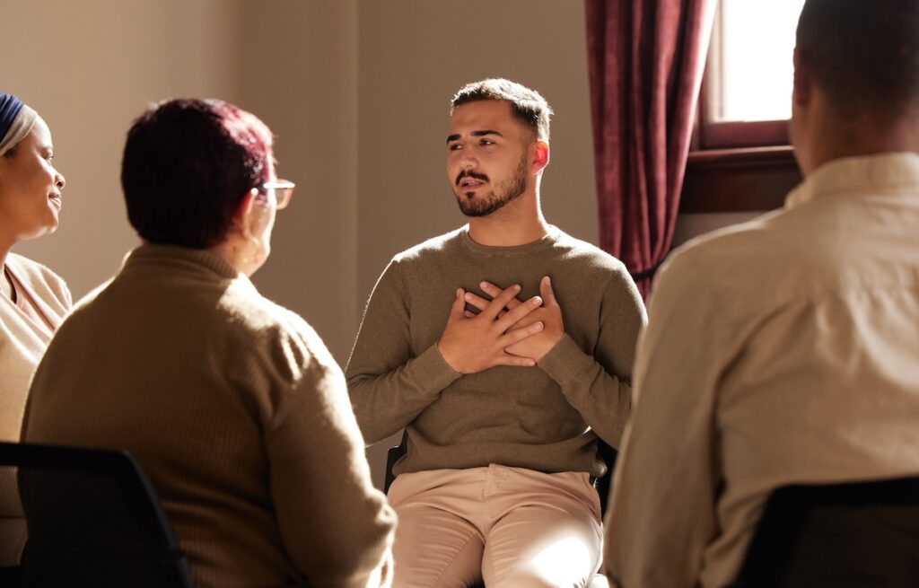 Man sitting in a group setting speaks with his hands on his chest, sharing his feelings while others listen attentively.