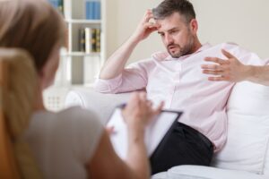 A man sits on a couch, looking distressed with one hand on his forehead while gesturing with the other, speaking to a therapist who is taking notes in the foreground.